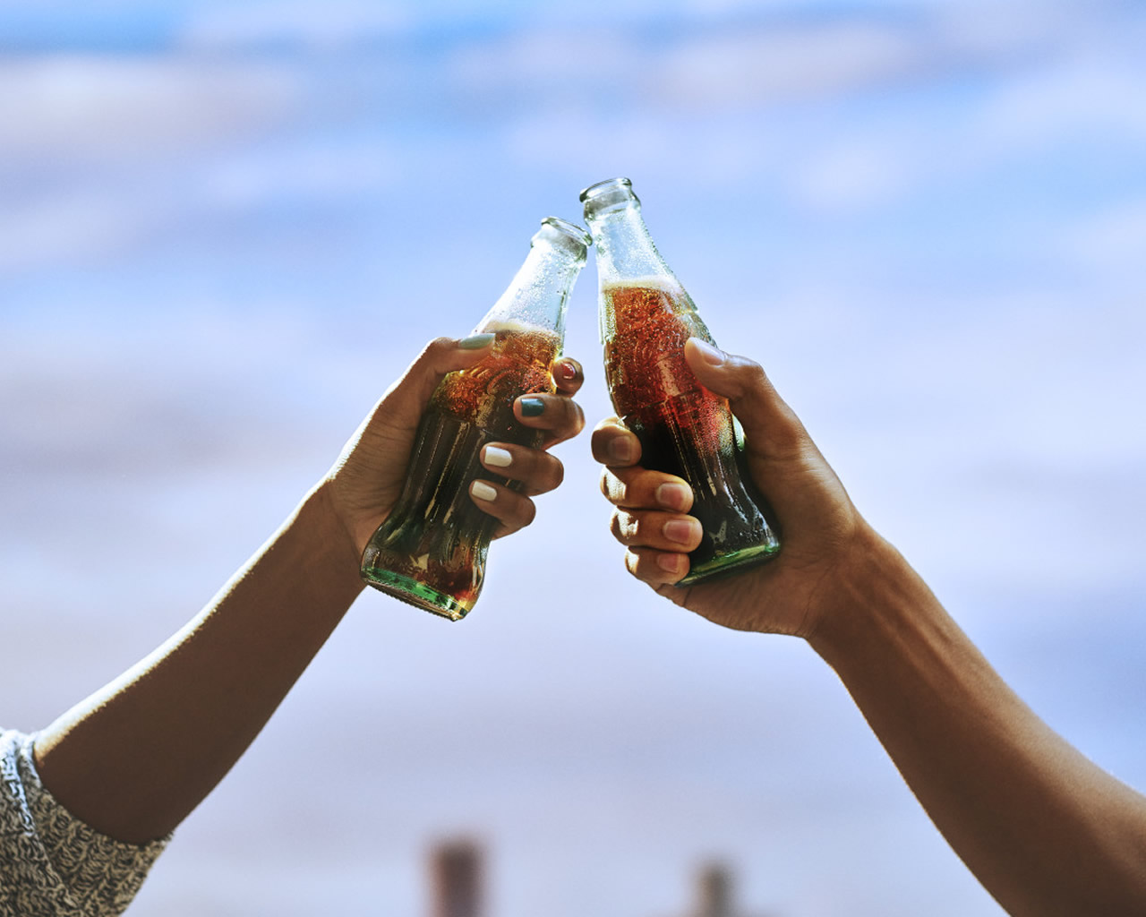 Zwei Hände mit Coca‑Cola Flaschen die anstossen