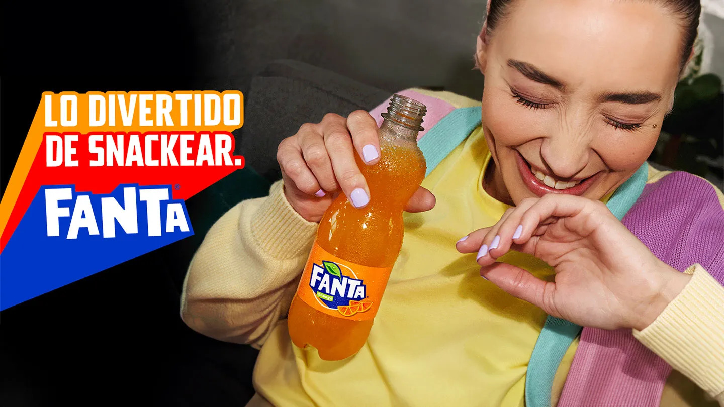 Mujer ríe divertida con una botella de Fanta naranja en la mano, a su izquierda hay un texto que dice “Lo divertido de snackear” y debajo el logo de Fanta.