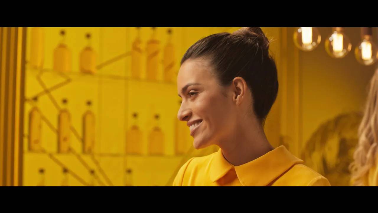 Γυναίκα με κίτρινο ρούχο χαμογελάει σε ένα μαγαζί με μπουκάλια και φώτα σε κίτρινο φόντο.