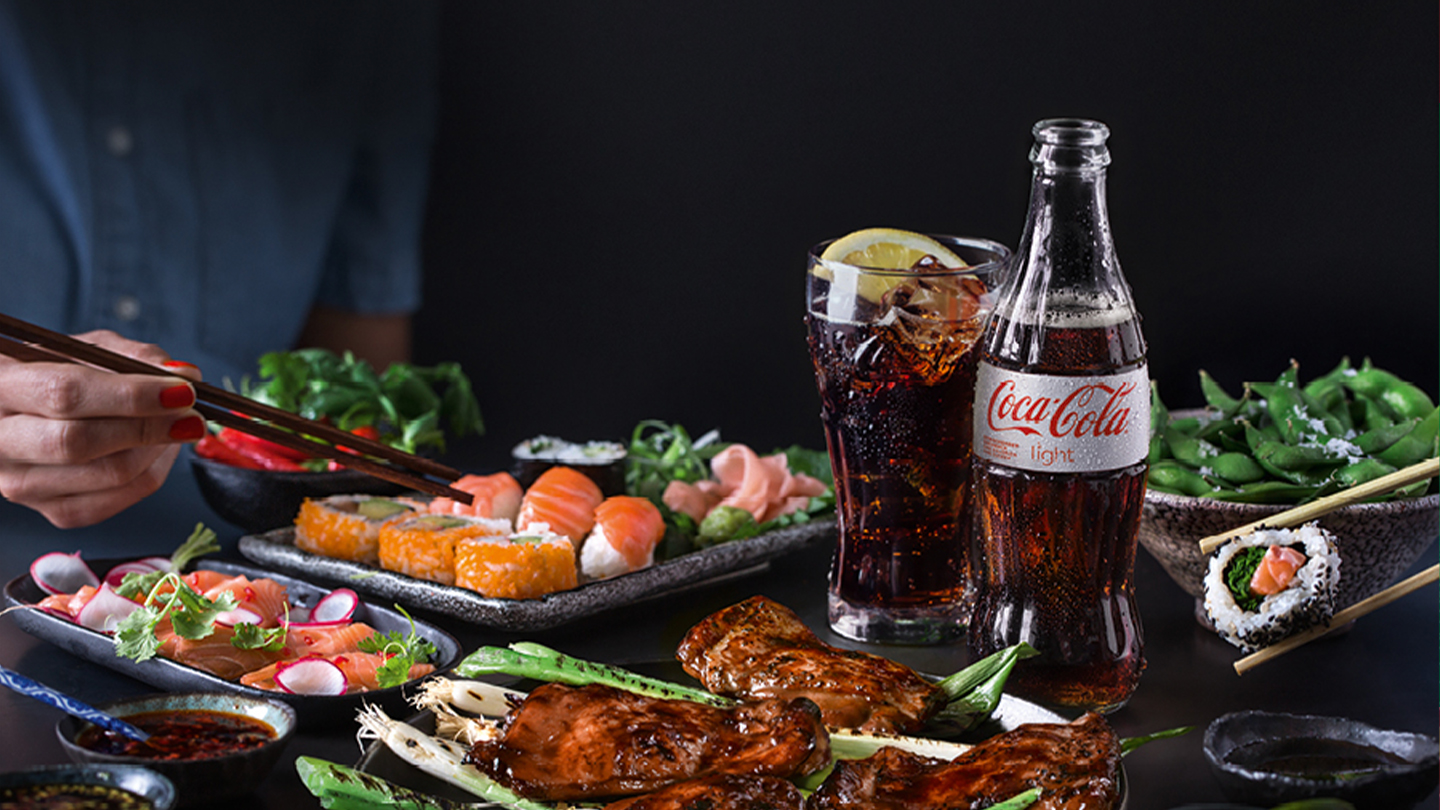 Mit Sushi und Gemüse Teller und Schüsseln auf einem Tisch, zwischen denen ein Glas mit Coca-Cola und eine Flasche Coca-Cola Light stehen. Links im Bild ist eine Hand mit Essstäbchen zu sehen.