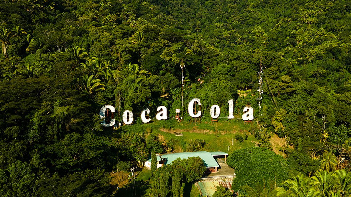 Stort Coca-Cola-skilt sammensat af individuelle bogstavsskilte på bakkeskråning blandt træer og huse