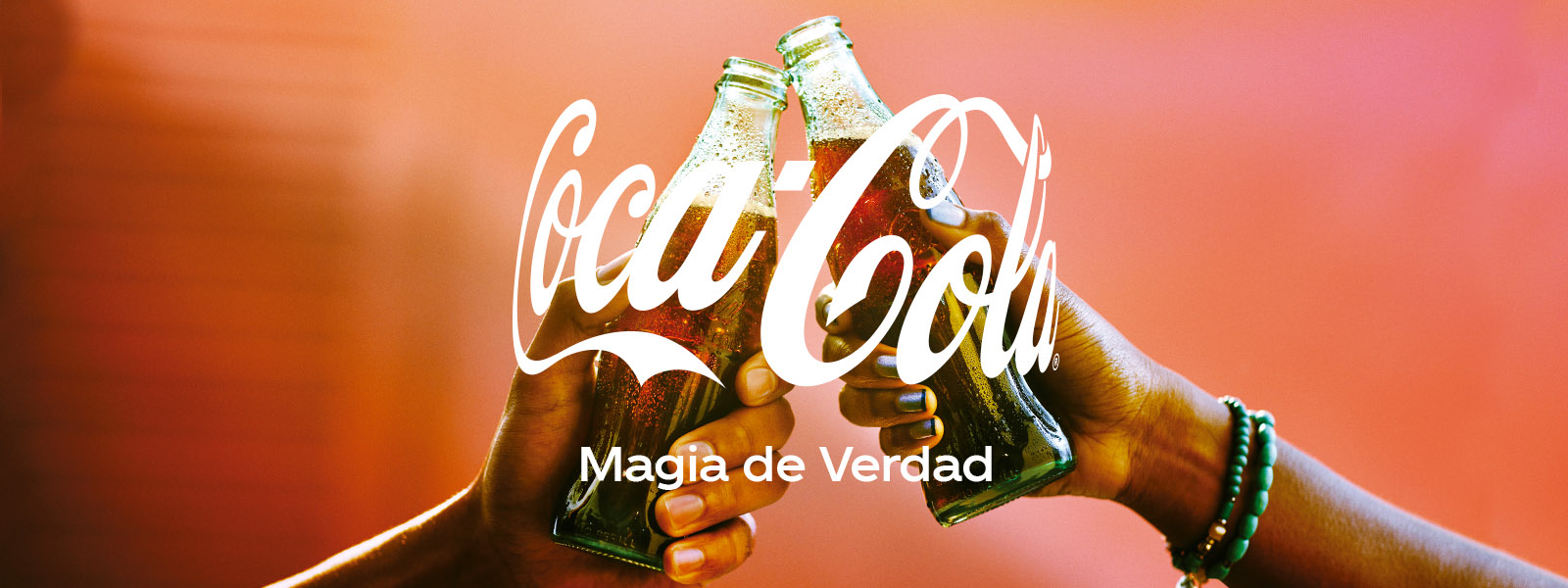 Dos manos sosteniendo una botella de vidrio de Coca-Cola brindando sobre un fondo difuso de color rojo. En el centro se observa el logo de Coca-Cola y el slogan “Magia de Verdad”.