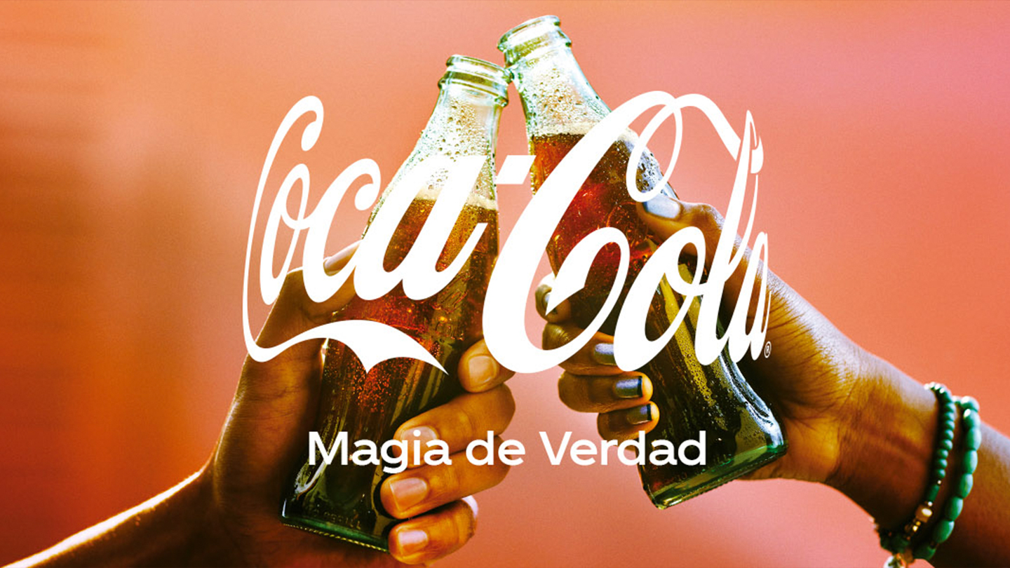 : Dos manos sosteniendo una botella de vidrio de Coca-Cola brindando sobre un fondo difuso de color rojo. En el centro se observa el logo de Coca-Cola y el slogan “Magia de Verdad”.
