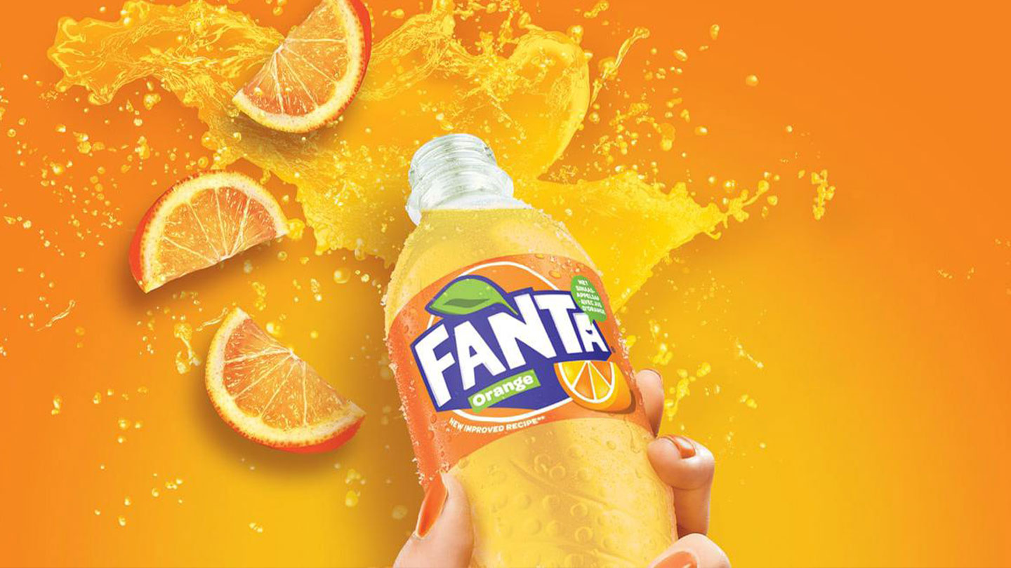 En Fanta-flaske hvor en spalsh av Fanta kommer ut og oppskjærete appelsiner ligger ved siden av