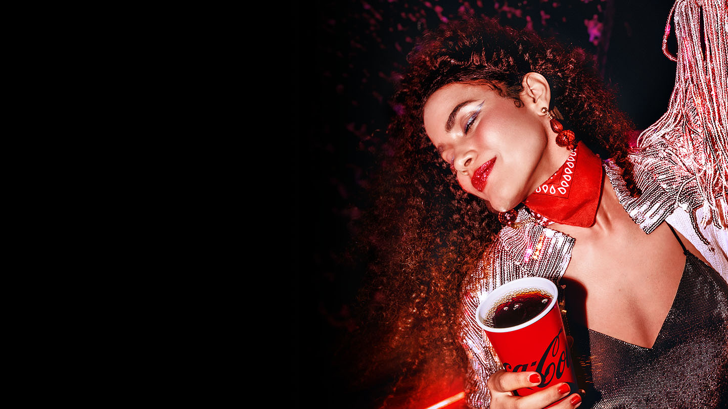Mujer disfrutando de una bebida Coca-Cola en un evento nocturno, luciendo un look brillante, labios rojos y una bandana roja, en un ambiente festivo y lleno de energía
