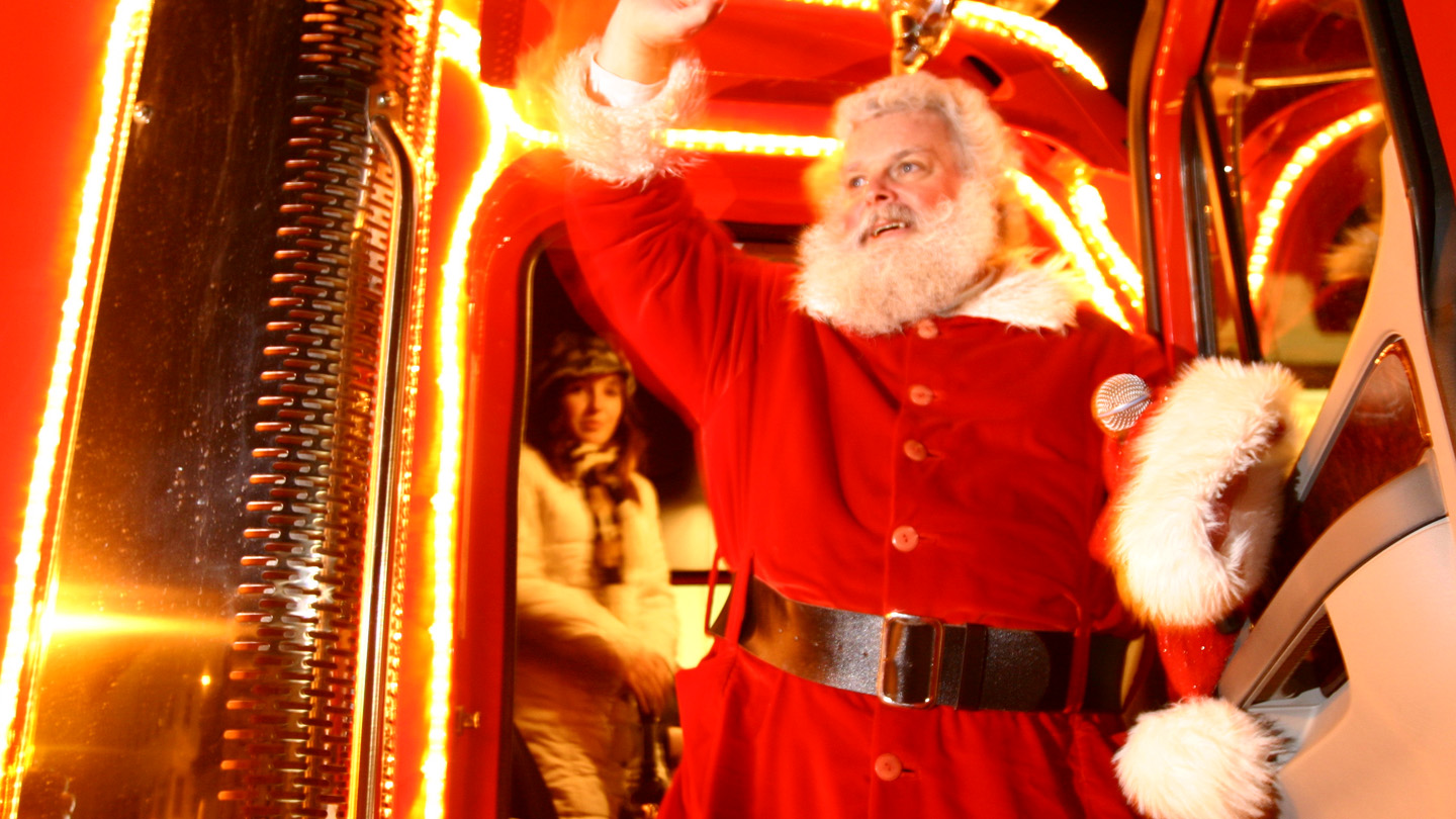 Der Coca-Cola Weihnachtsmann steht winkend vor dem Coca-Cola Weihnachtstruck