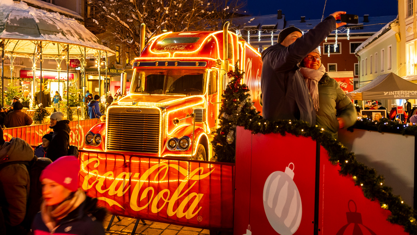 Coca-Cola Weihnachtstruck, leuchtend rot, umgeben von Zuschauern bei Dämmerung auf einem Platz