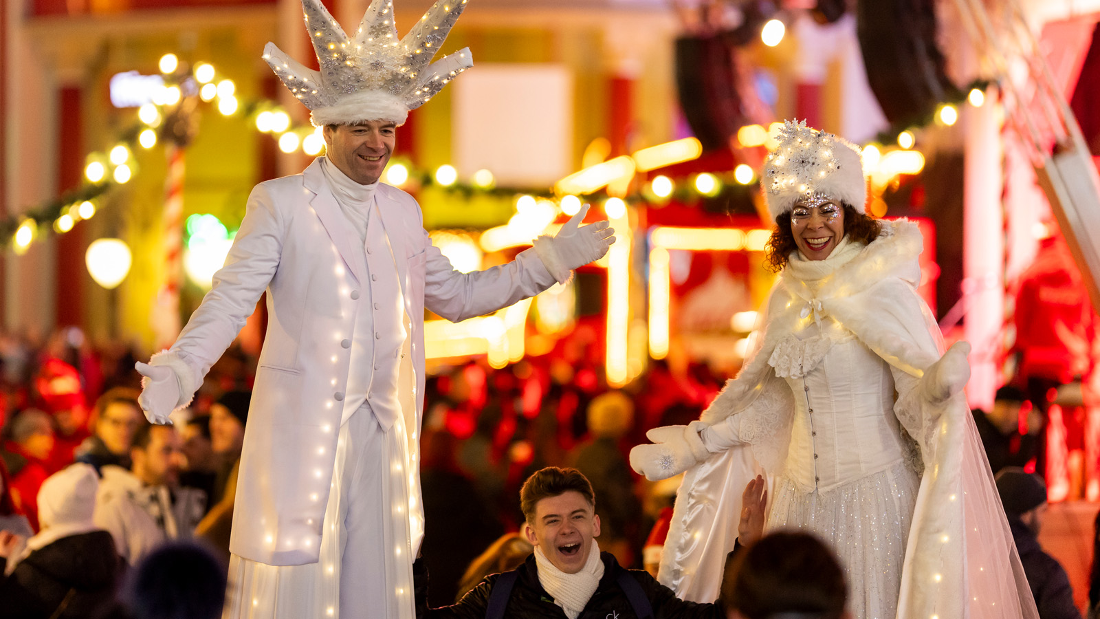 Zwei leuchtende Figuren auf Stelzen auf einem Weihnachtsmarkt, mit einem begeisterten Mann zwischen ihnen, umgeben von Menschen.