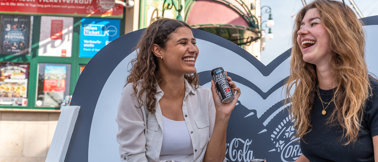 Zwei Personen sitzen lachend mit einem Coca-Cola OREO auf einer Bestie Bank