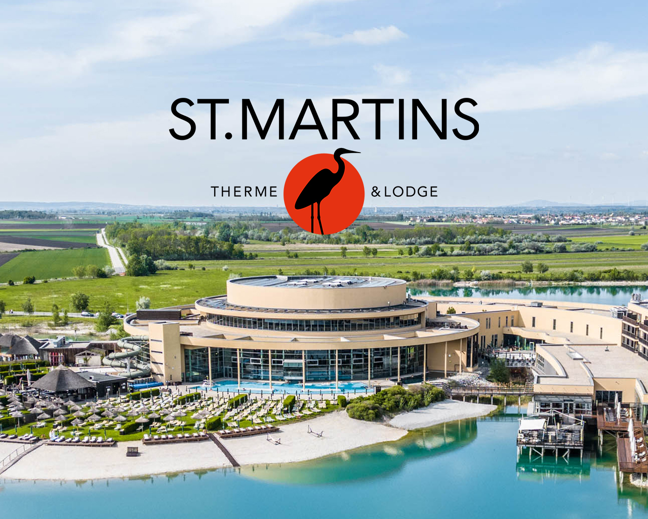 Luftaufnahme der St. Martins Therme & Lodge mit dem Thermengebäude, Liegewiese, Rutsche und angrenzendem See. Darüber das Logo mit dem Schriftzug „St. Martins Therme & Lodge" und einem Reiher vor roter Kreisfläche.