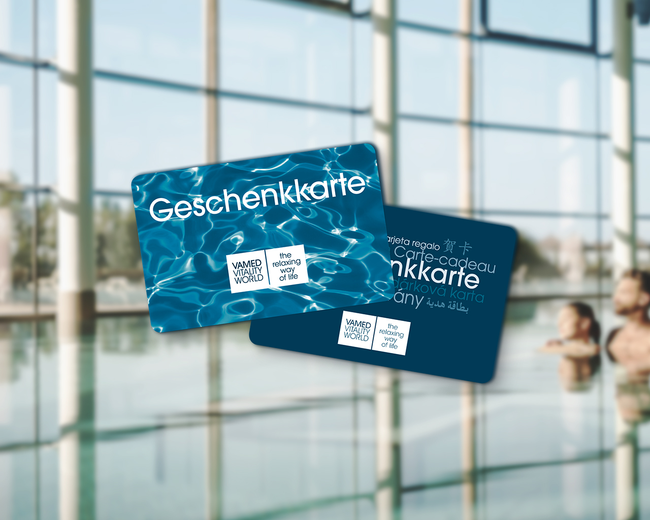 Zwei Geschenkkarten der Vamed Vitality World vor dem verschwommenen Hintergrund einer Therme mit Glasfront und Wasserbecken. Die vordere Karte zeigt ein Wassermuster und den Schriftzug „Geschenkkarte", die hintere die Bezeichnung „Geschenkkarte" in mehreren Sprachen.