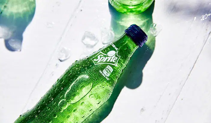 A Sprite bottle on top of a white surface with a few pieces of ice close to it