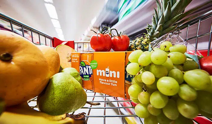 A view from inside of a supermarket cart where there are various types of fruit and a Fanta Orange packaging in the middle