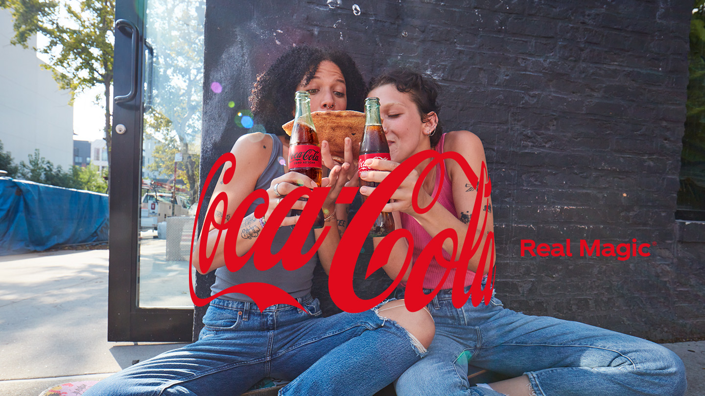 Dvoje prijatelja sjede na podu i uživaju u boci Coca-Cole i komadu pizze.