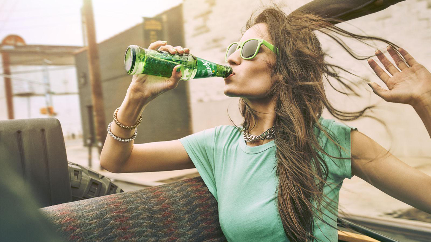 Mlada žena uživa u ispijanju Spritea iz boce noseći zelenu majicu i sunčane naočale