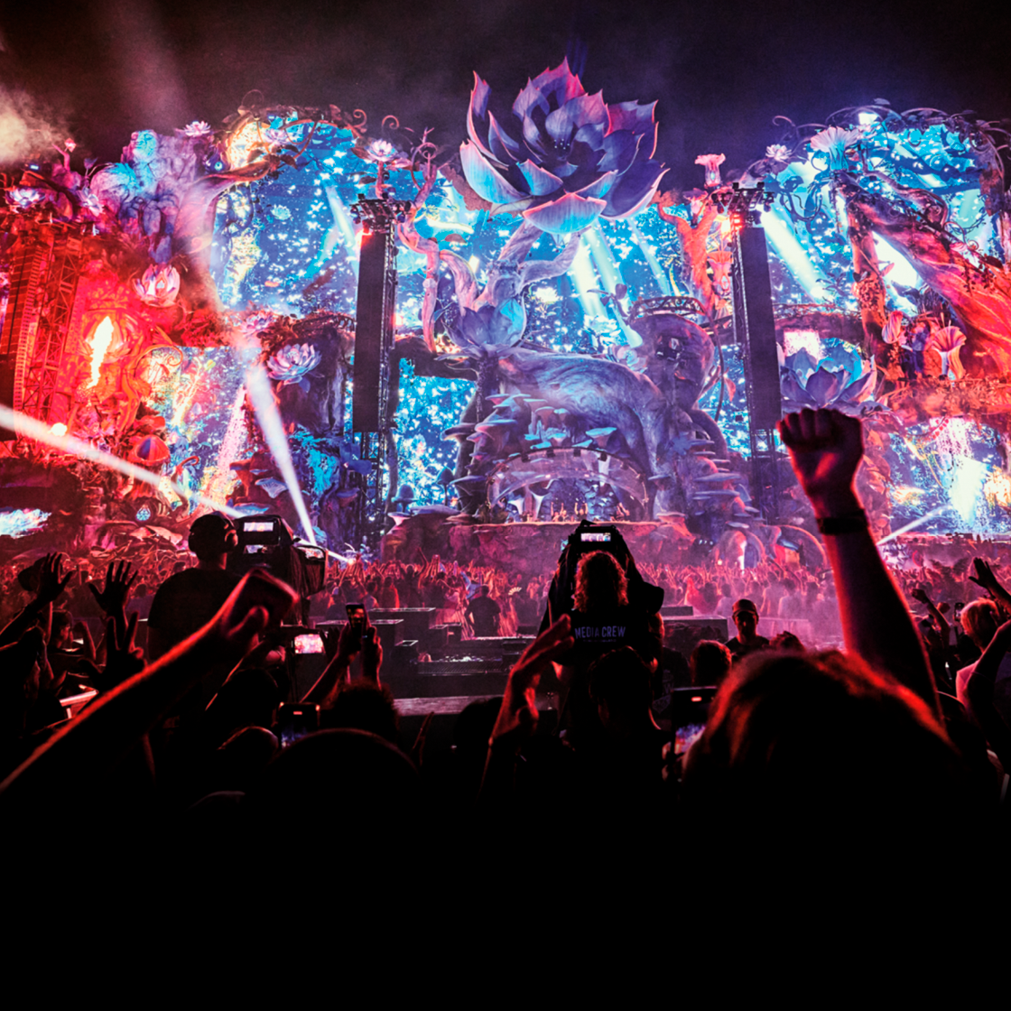 Une grande scène de festival de musique, colorée, avec des décorations fantaisistes, des lumières vives et des effets visuels, vue depuis le public. Au premier plan, des personnes lèvent les bras et leurs téléphones pour filmer le spectacle.