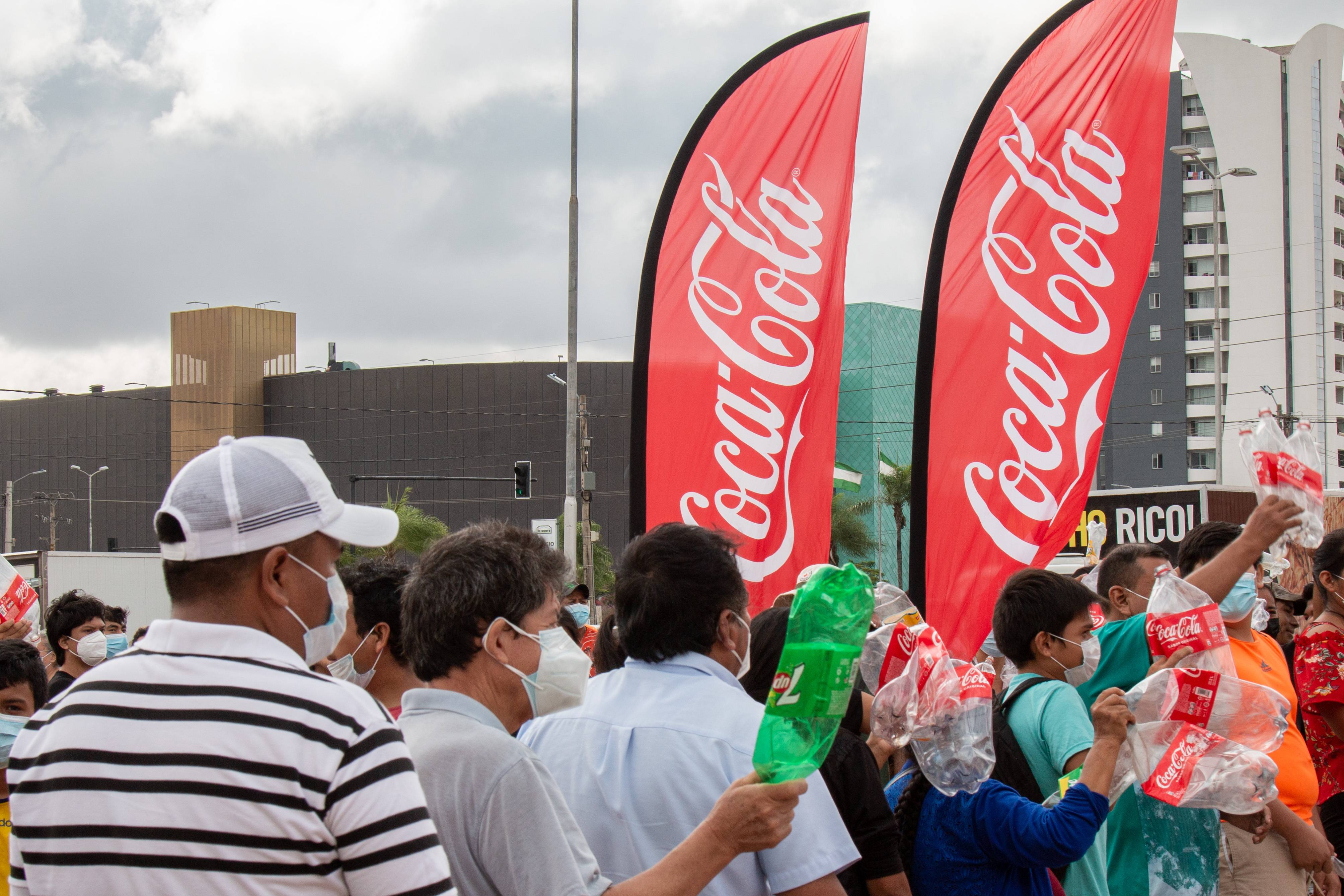 2 banderas de Coca-Cola y personas con botellas plásticas en la mano