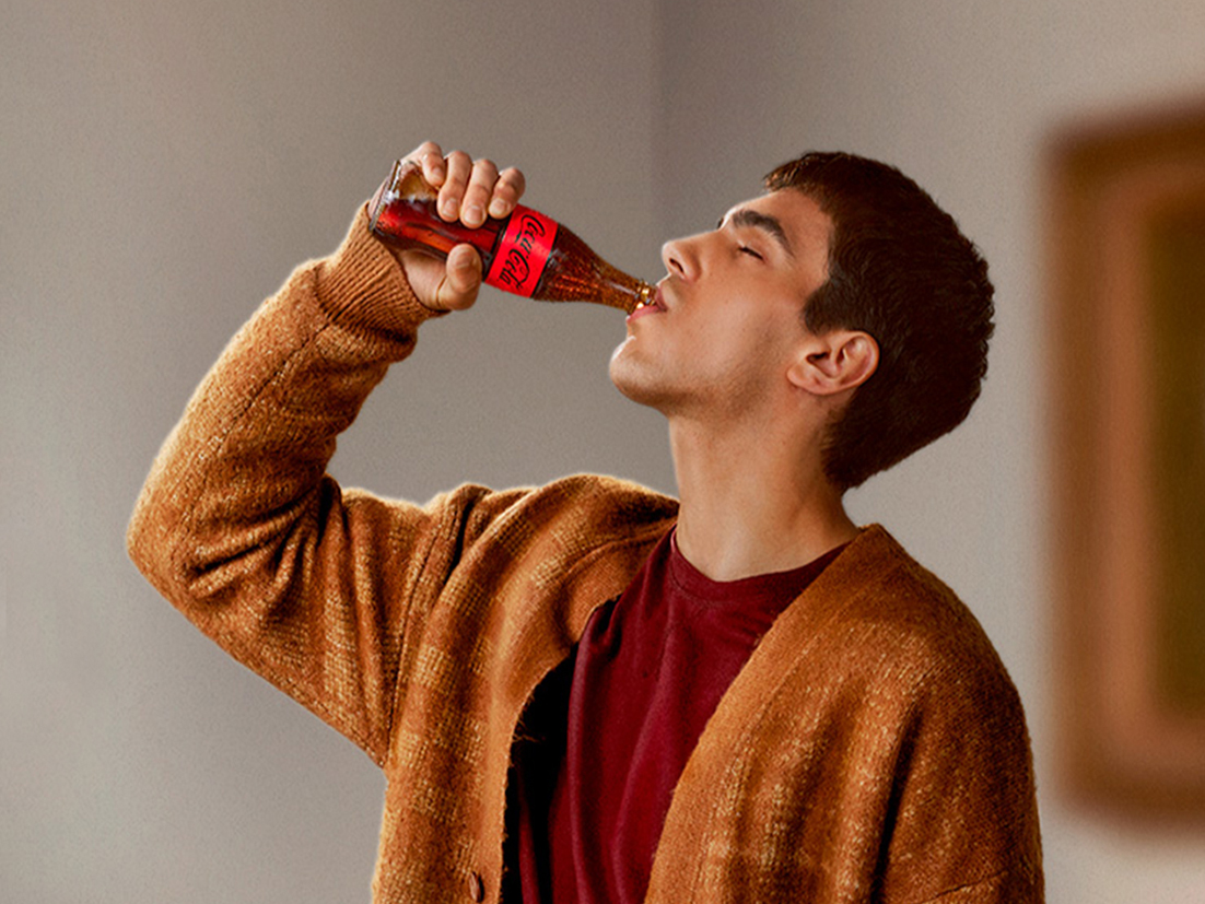 Hombre de pelo corto y vestimenta formal bebiendo una botella de Coca-Cola de vidrio con los ojos cerrados en un museo.
