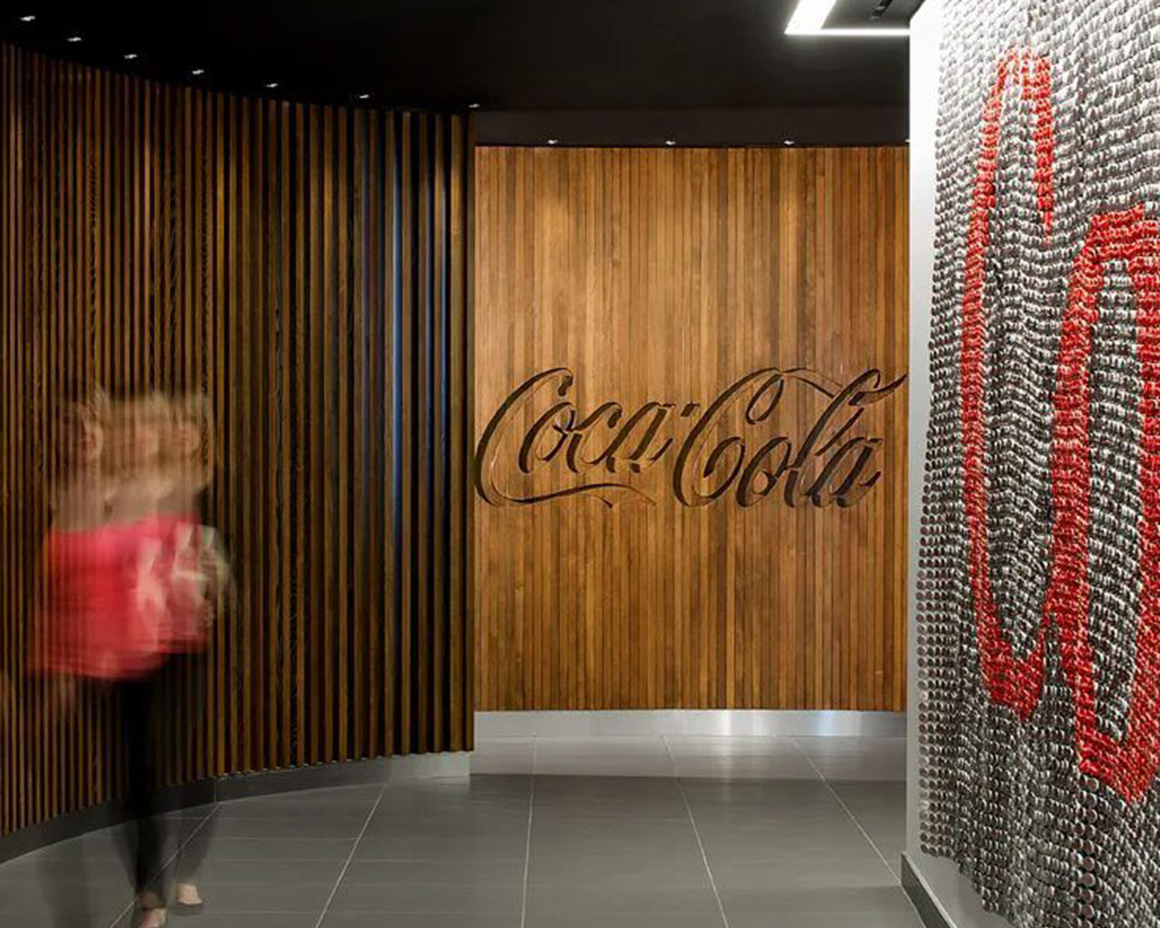 Mujer rubia caminando por una oficina de paredes de madera donde se observa el logo de Coca-Cola en la pared del fondo.