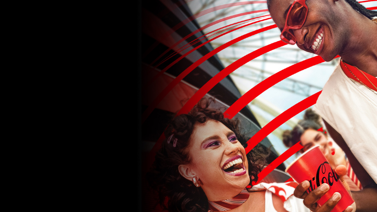 Dois amigos sorrindo alegremente enquanto seguram copos vermelhos da Coca-Cola. O fundo mostra um ambiente ao ar livre com uma estrutura metálica e efeitos gráficos de linhas vermelhas em destaque.