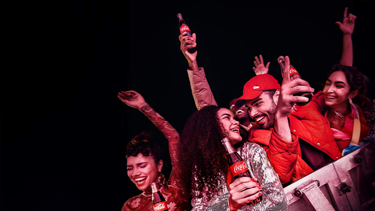 Jovens sorrindo e celebrando em um ambiente escuro, alguns segurando garrafas de Coca-Cola.