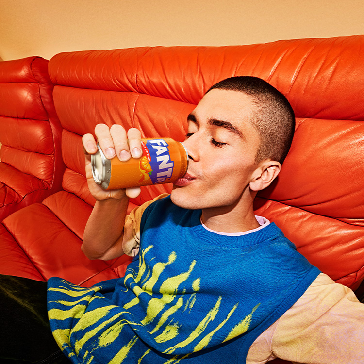 Um jovem está deitado em um sofá de couro laranja vibrante, bebendo uma lata de Fanta. Ele está vestindo uma camiseta azul com um desenho amarelo.