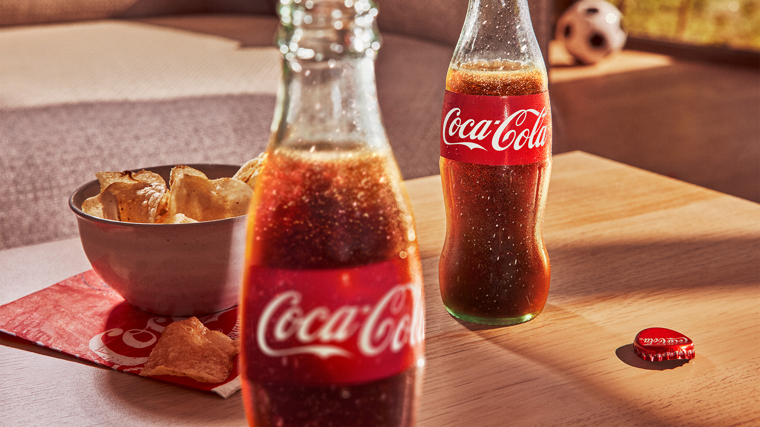 Two glass bottles of Coca-Cola sit on a wooden table beside a bowl of chips, a Coca-Cola bottle cap, and a soccer ball visible in the background.