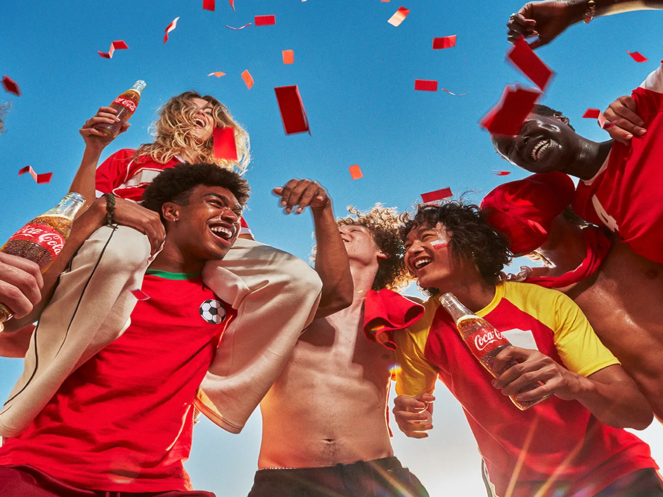 Soccer fans in red jerseys celebrating together, holding Coca‑Cola bottles as red confetti falls against a bright blue sky.