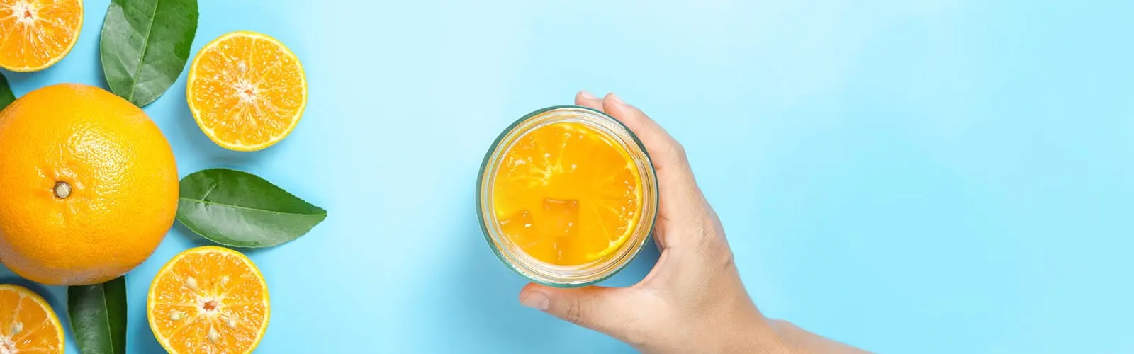 Top view of an orange, two oranges cut in half, and a hand holding a glass of orange juice with ice on a light blue surface