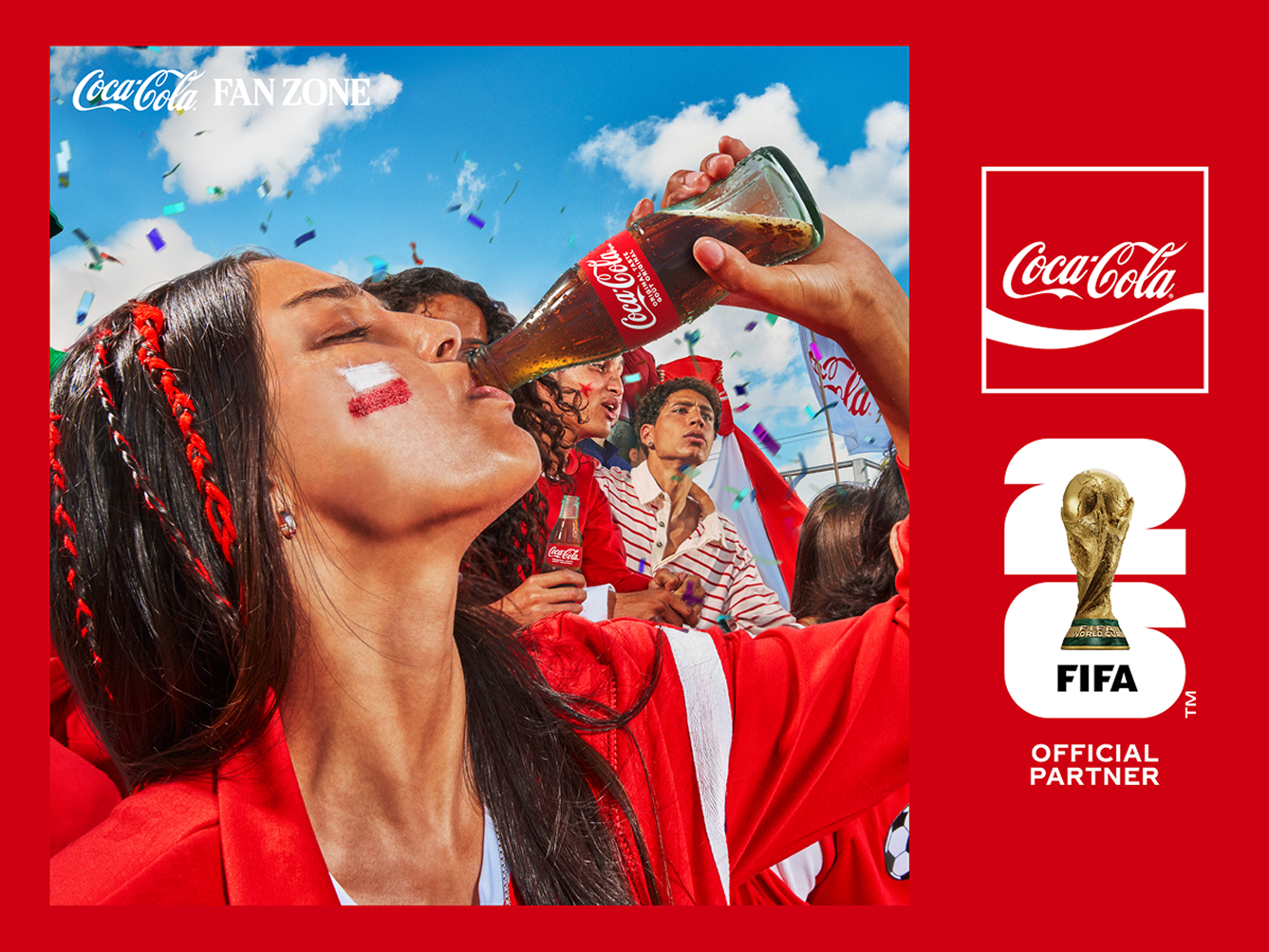 A young soccer fan with face paint drinks from a classic Coca-Cola bottle while celebrating with a crowd under a blue sky at a Fan Zone.