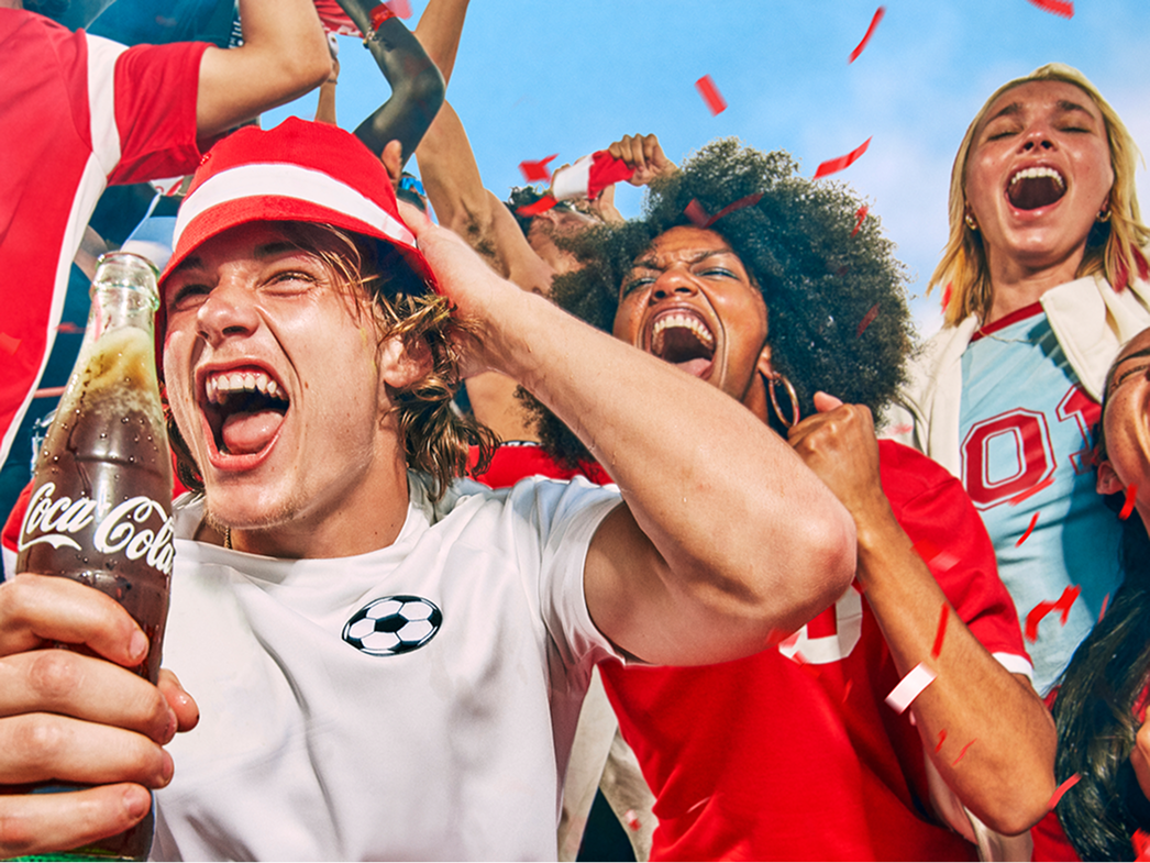 Group of soccer fans in stands celebrating a goal, tossing red confetti while one fan holds a chilled Coca-Cola bottle in foreground.