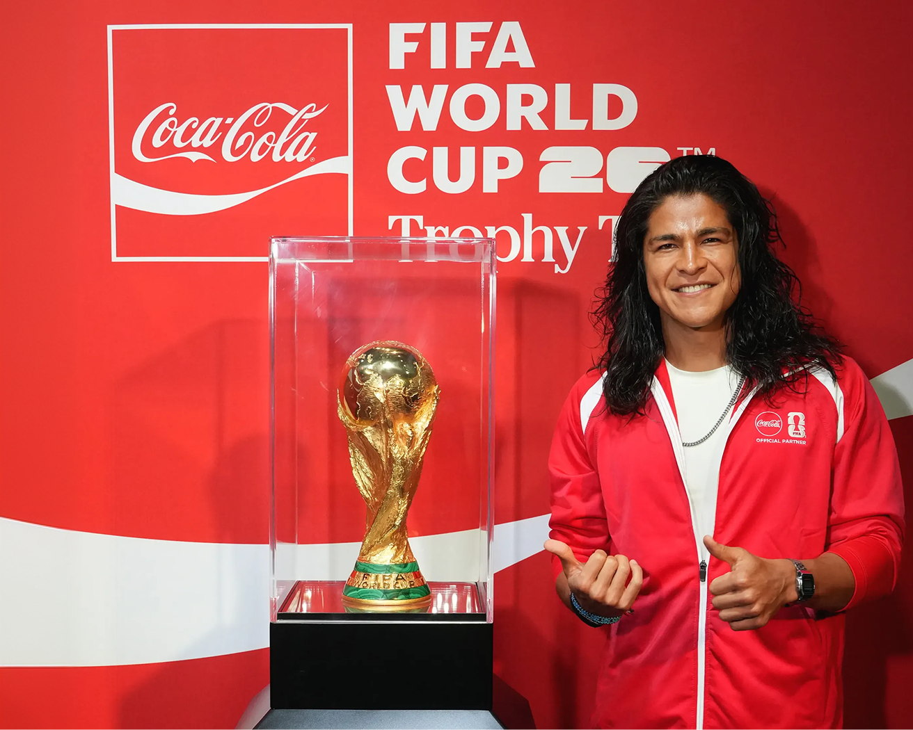 Fan in red Coca-Cola jacket posing beside FIFA World Cup original trophy in glass case at Trophy Tour event desktop image