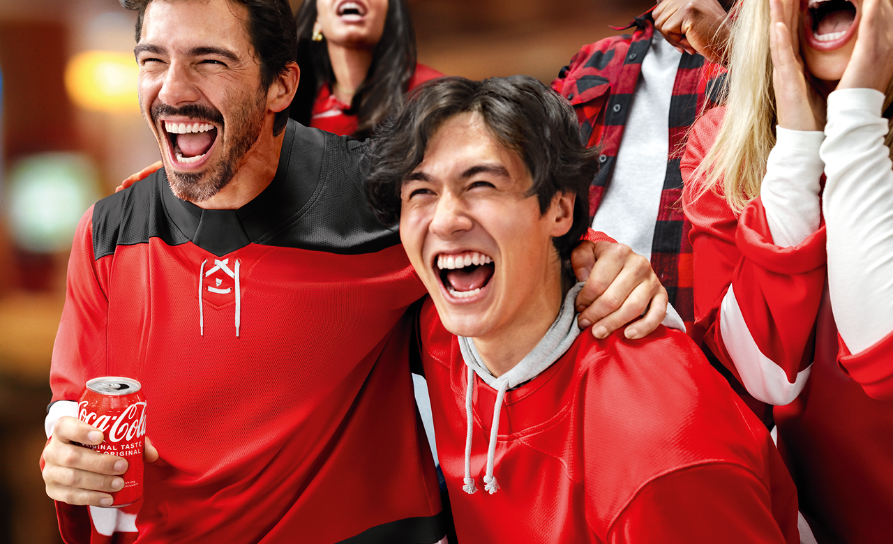 Excited hockey fans in red jerseys cheer in the arena as one holds an ice‑cold Coca‑Cola can during a fall game.