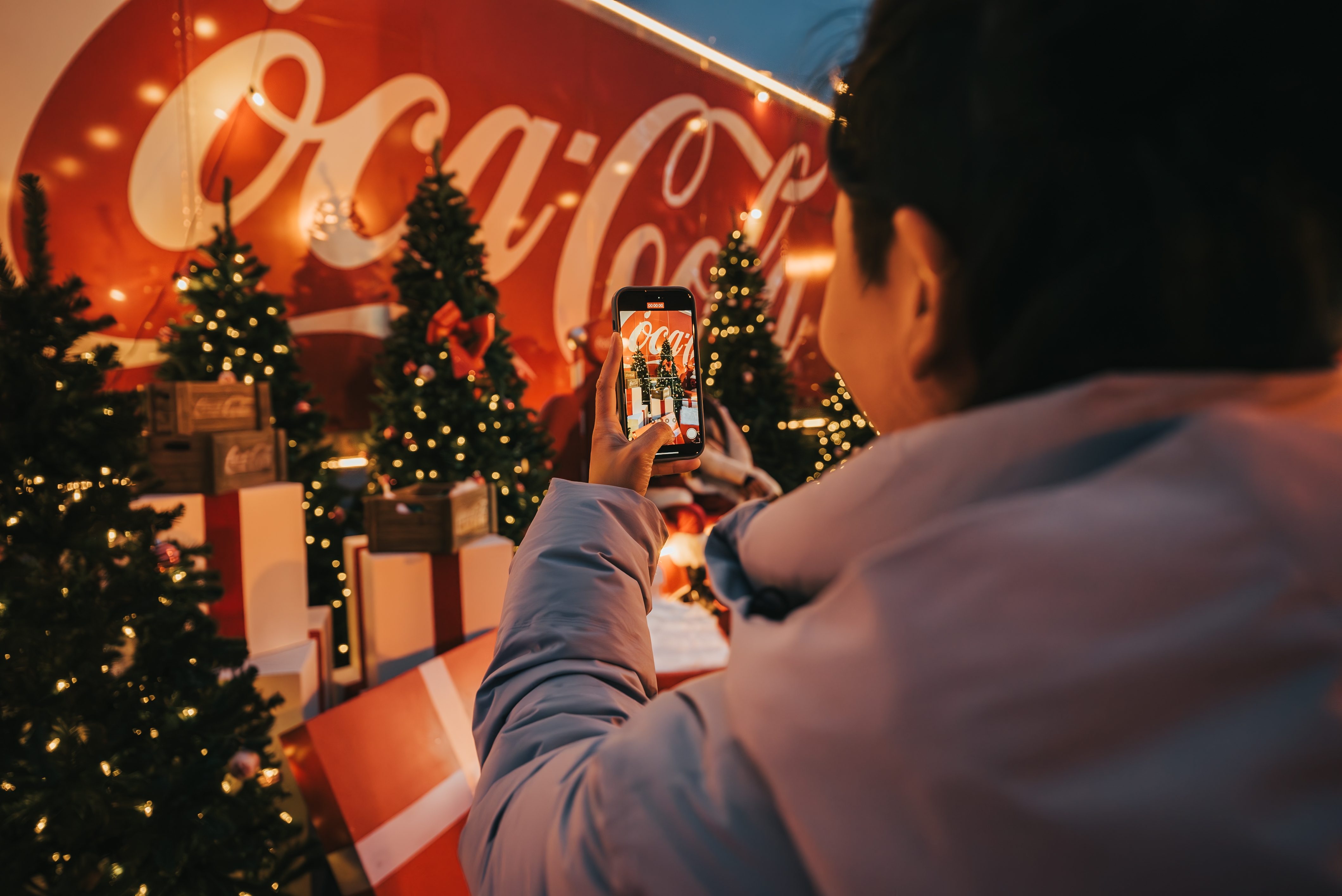 Par-dessus l'épaule, une personne photographie une scène festive avec son téléphone intelligent : des sapins de Noël illuminés et des cadeaux sont placés devant le camion rouge du Caravan des Fêtes Coca-Cola.