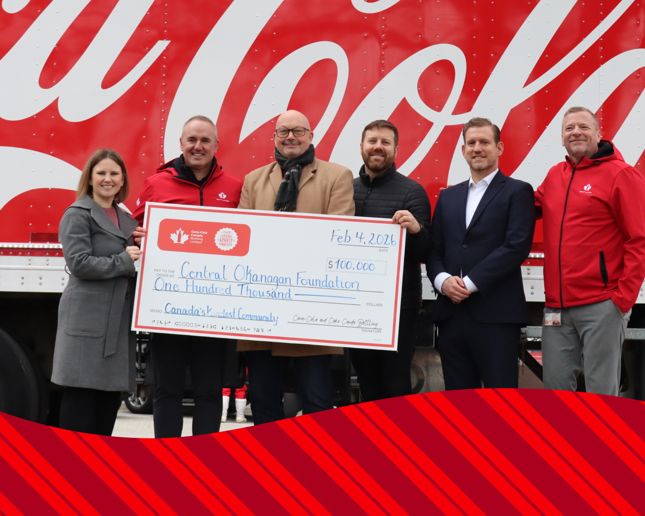 Personnes se tiennent devant un camion de livraison Coca-Cola en tenant un chèque géant de 100 000 $ daté du 4 février 2026, payable à la Fondation Central Okanagan, avec la mention « Canada's Kindest Community », entourées de motifs d'ornements de Noël.