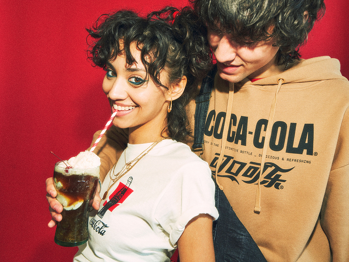 Une jeune femme buvant un flotteur au cola avec une paille rayée se tient devant un jeune homme portant un chandail à capuchon beige imprimé du logo « Coca-Cola », sur fond rouge.