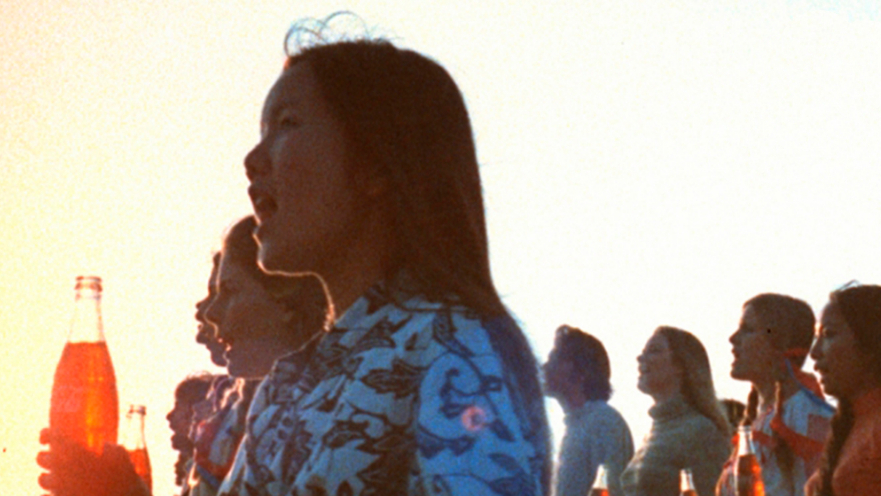 A girl holding a coke bootle and sings with a group of people. Image from the music video of the song Hilltop.