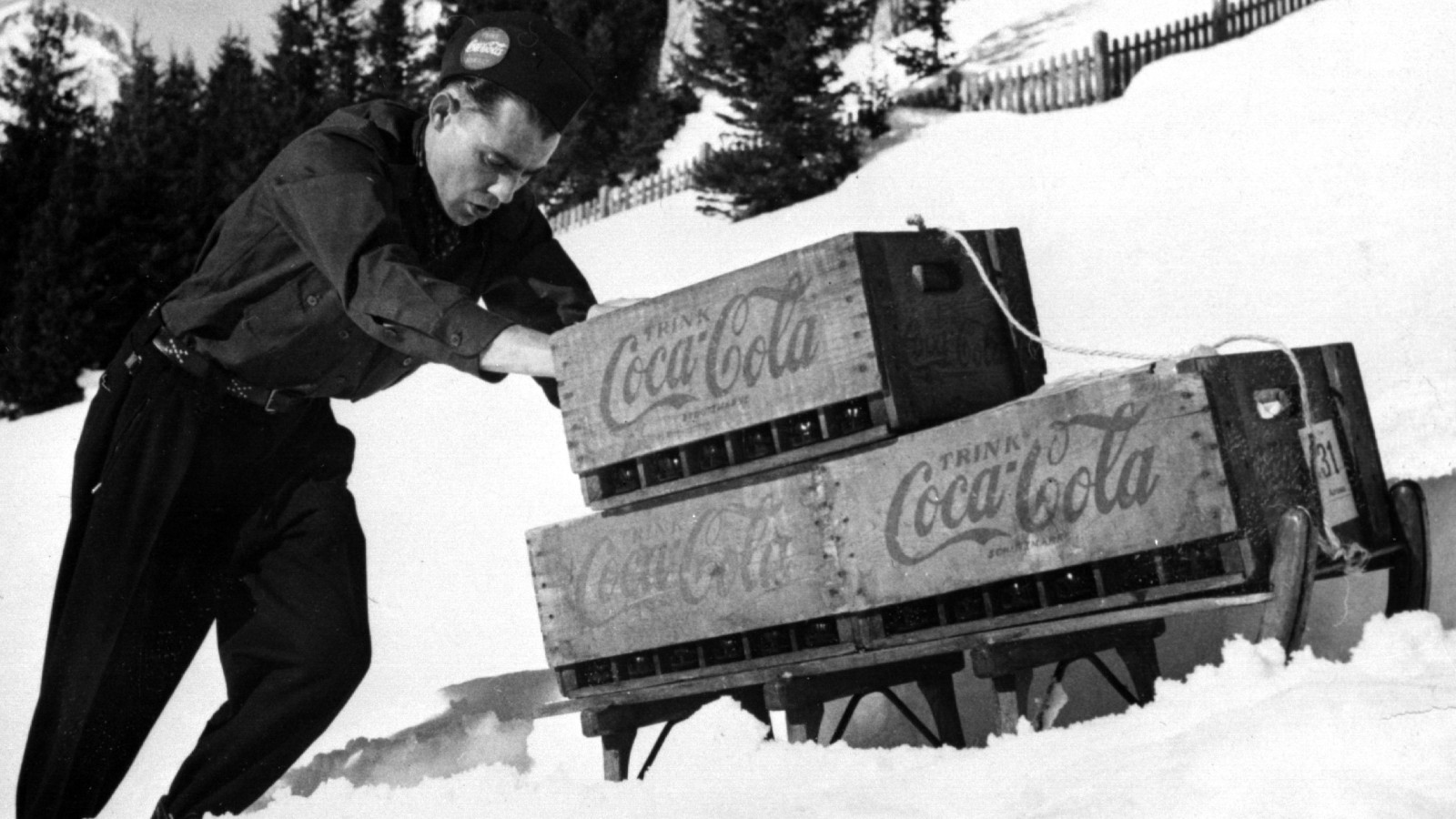 Photo historique d’un homme dans la neige avec une luge chargée de caisses de Coca-Cola.