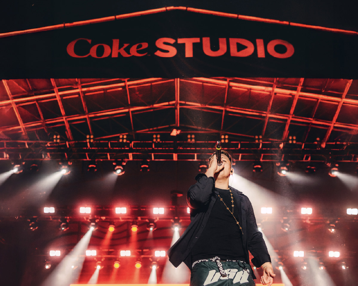 Un artista actuando en el escenario principal del festival Coke Studio, bajo una estructura iluminada con luces rojas y blancas. En la parte superior, se destaca el logotipo de "Coke Studio" en letras grandes y brillantes, creando un ambiente vibrante y lleno de energía.