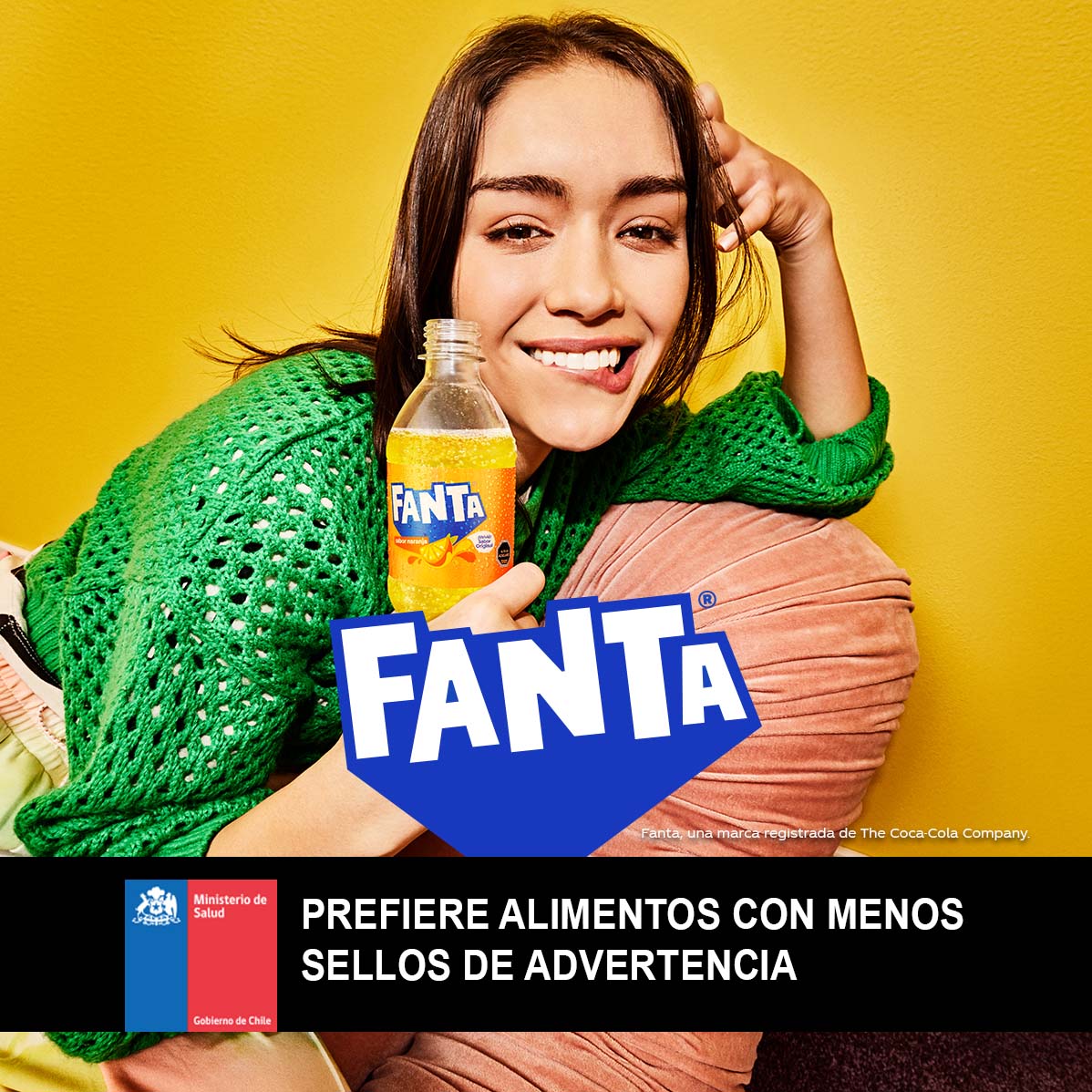 Una joven sonríe con una botella de Fanta Naranja, vestida con un suéter verde y recostada sobre un almohadón en una habitación amarilla.