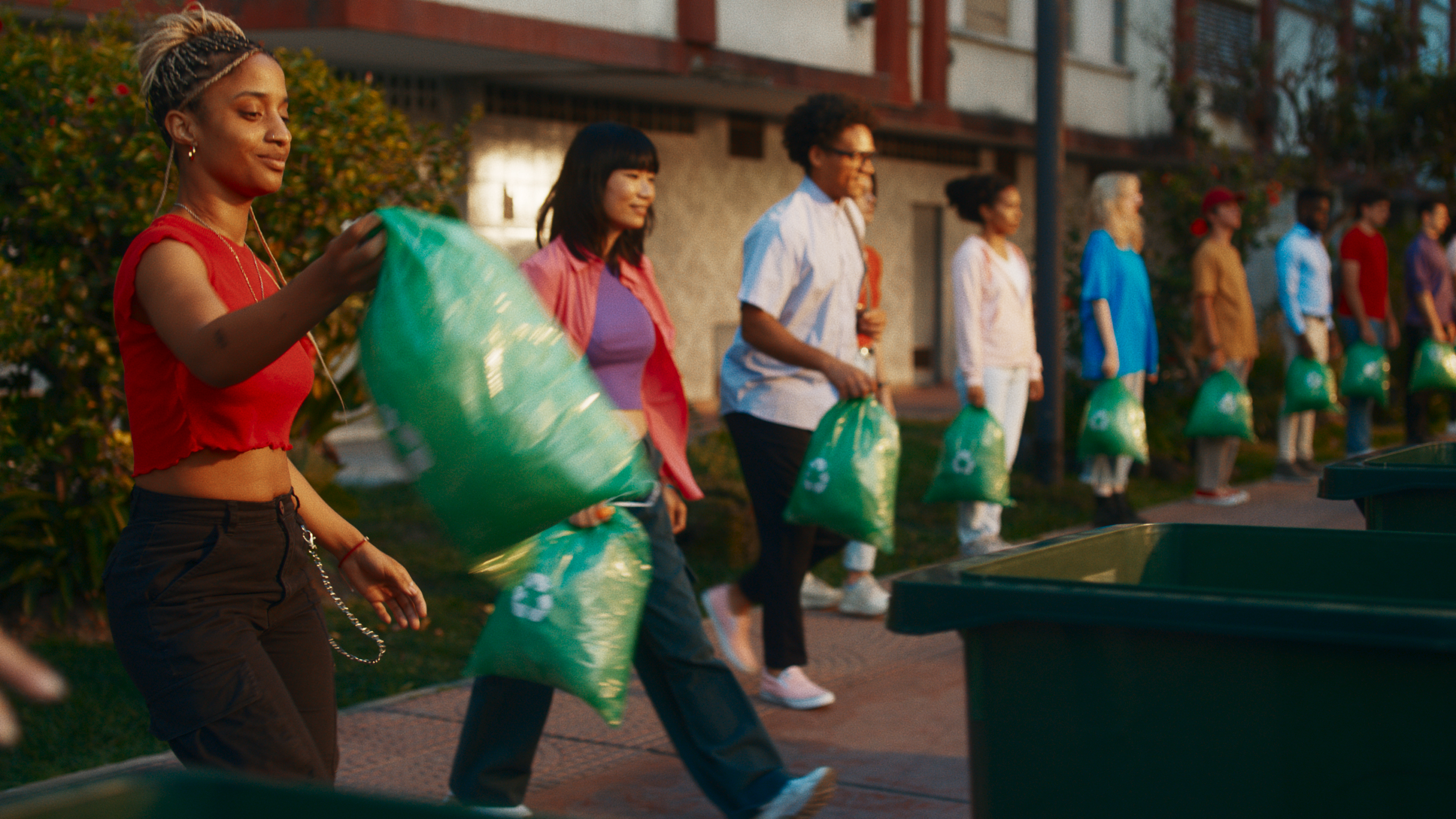 Hombres y mujeres en fila con bolsas verdes en sus manos.  La mujer en primer plano lleva su bolsa a un basurero.