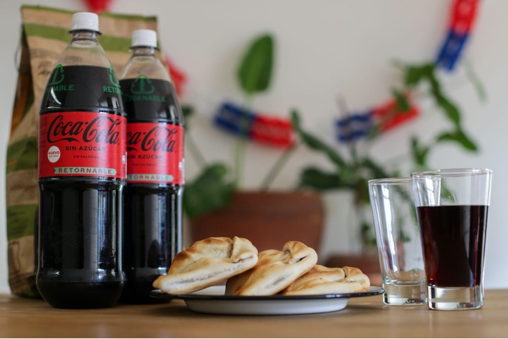 Dos botellas de Coca-Cola junto a plato con 3 empanadas y dos vasos, uno vacío y el otro lleno.