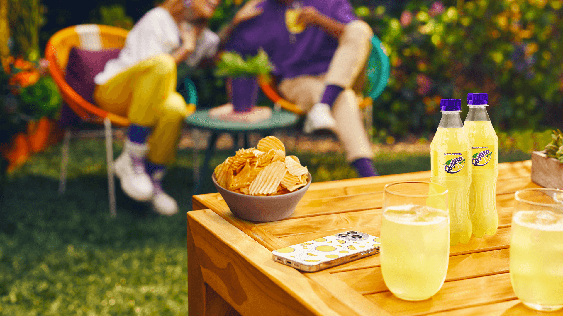 Escena de almuerzo al aire libre. Una mesa de madera con botellas de Quatro y vasos con hielo y gaseosa junto a unas papas fritas y un teléfono celular. De fondo y fuera de foco, dos personas sentadas en sillas de jardín conversando.