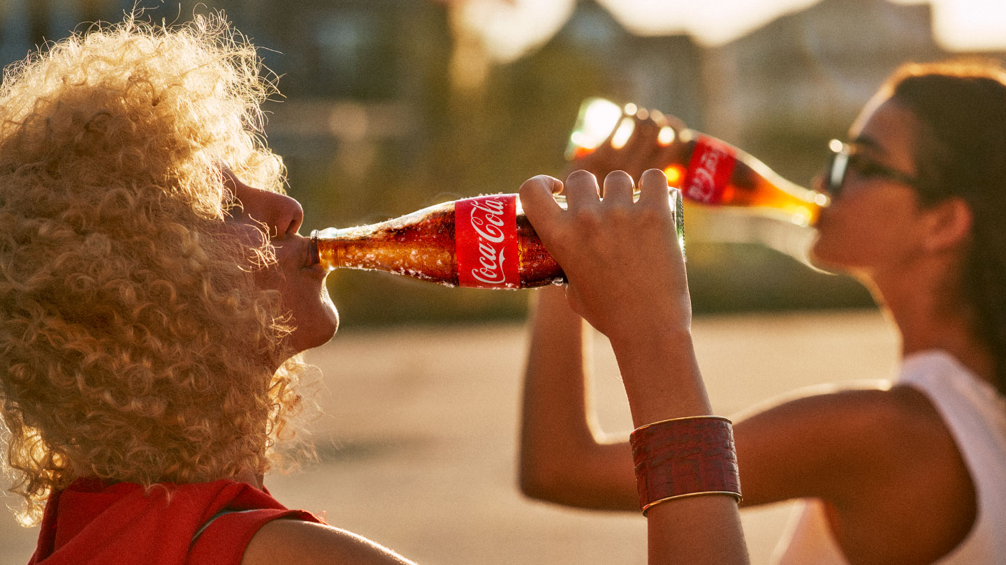 Mujer rubia de pelo enrulado bebiendo una botella de Coca-Cola de vidrio al aire libre en el atardecer