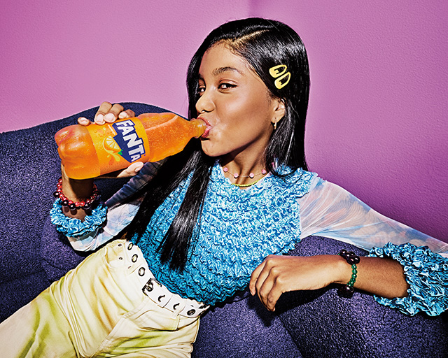 Mujer bebiendo de una botella de Fanta Naranja recostada en un sillon.