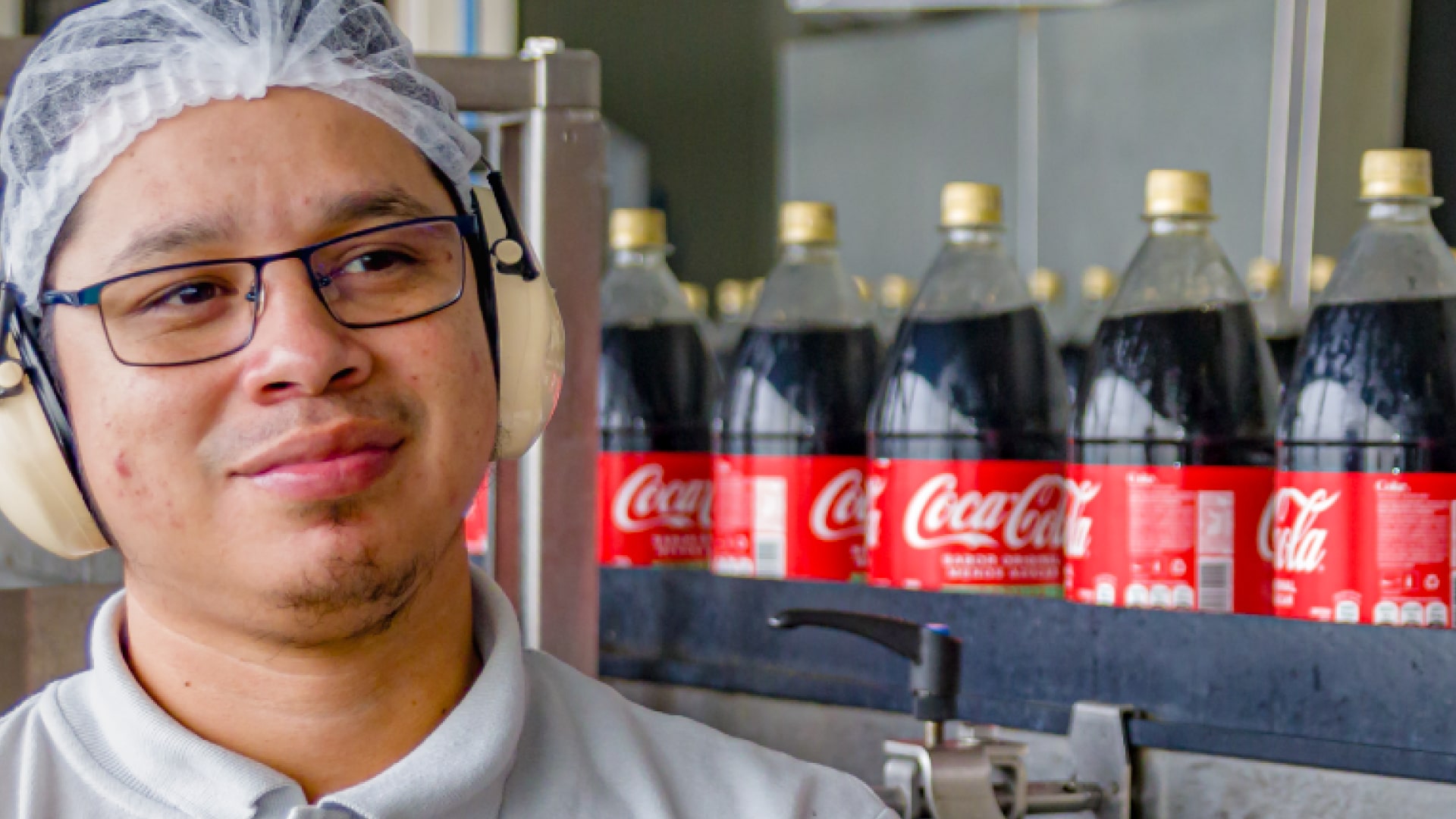  Un hombre joven con gafas, una redecilla blanca en la cabeza y protectores auditivos de color crema, sonríe ligeramente mientras mira hacia la izquierda. Detrás de él, se ven varias botellas de Coca-Cola en una línea de producción, con sus etiquetas rojas y tapas amarillas claramente visibles.
