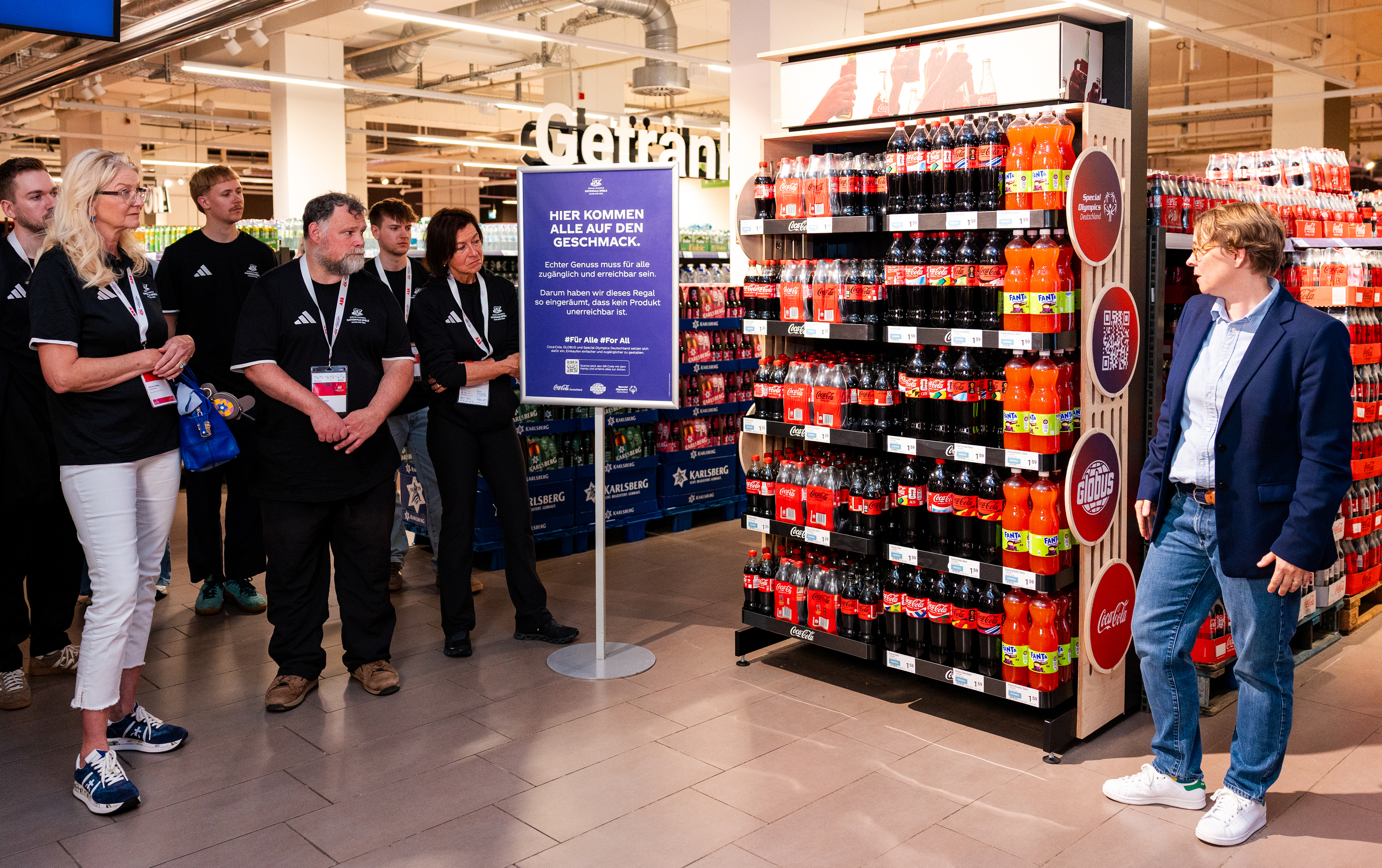 Gruppe von Teilnehmenden bei einer Marktbegehung in einer GLOBUS-Markthalle vor einem Getränkeregal mit Coca-Cola Produkten und Hinweisschild zu zugänglicherem Einkaufen.