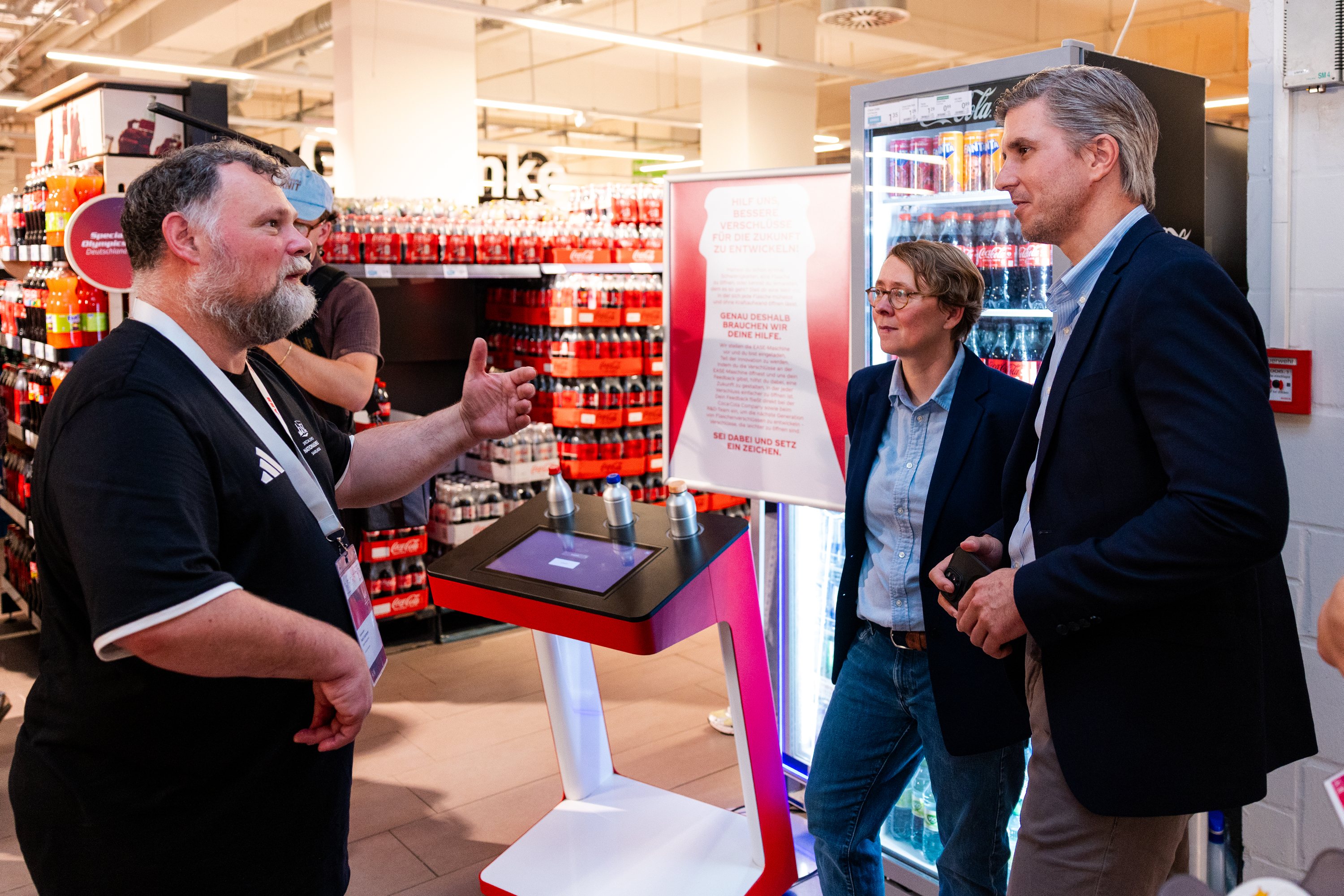 Teilnehmende des Pilotprojekts von Coca-Cola, GLOBUS und Special Olympics Deutschland im Gespräch an einer Teststation für leichter zu öffnende Verschlüsse in einer GLOBUS-Markthalle.