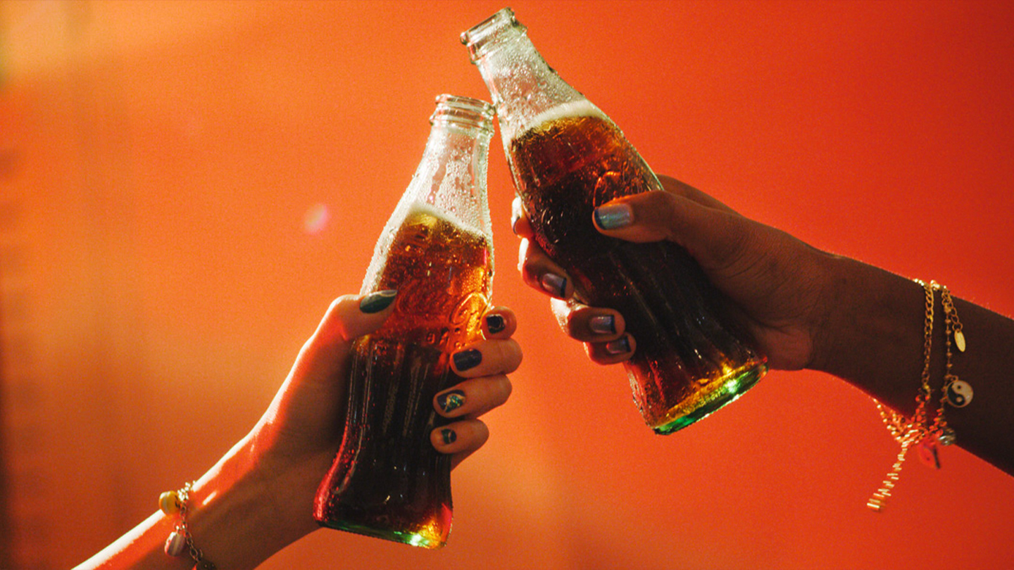 Zwei Hände halten je eine gläserne Flasche Coca-Cola und stoßen damit an.