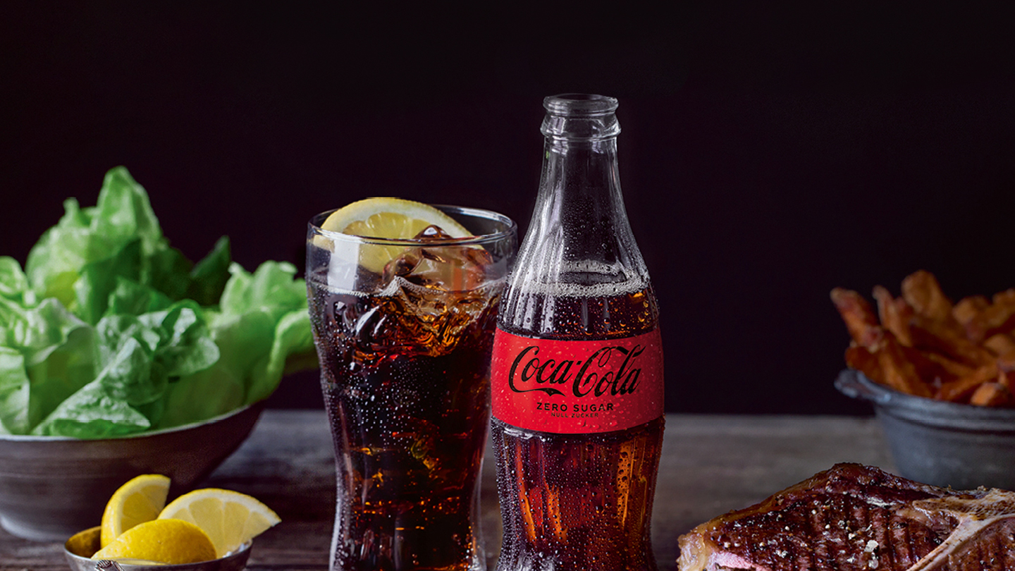Eine Flasche Coca-Cola Zero Sugar und ein gefülltes Glas auf einem Tisch mit verschiedenen Speisen